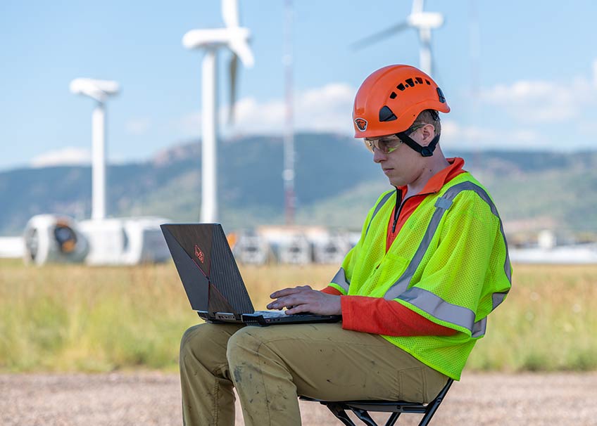 A researcher in a yellow safety vest and orange helmet types on a laptop outside with wind turbines in the distance. 