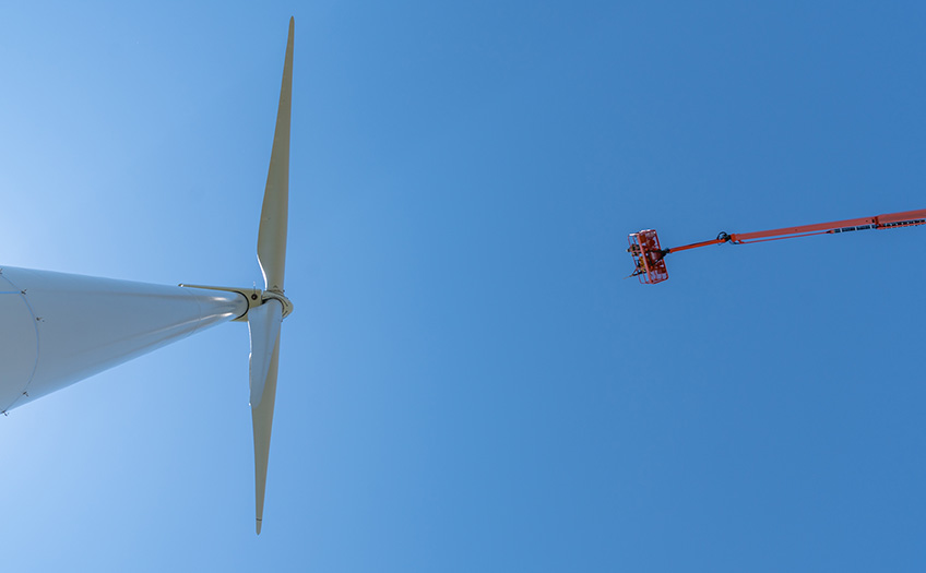 Two men on an orange manlift launch tennis balls at a wind turbine from a potato cannon.