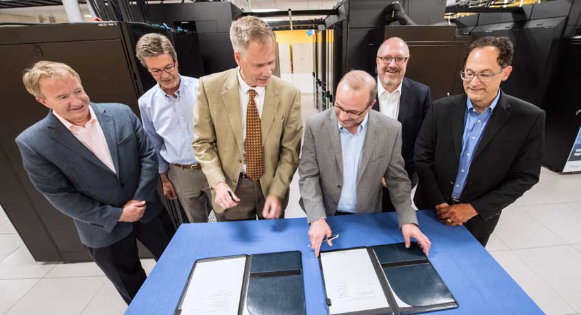 Photo of a group of men standing around a table in a data center. Two men sign pieces of paper.