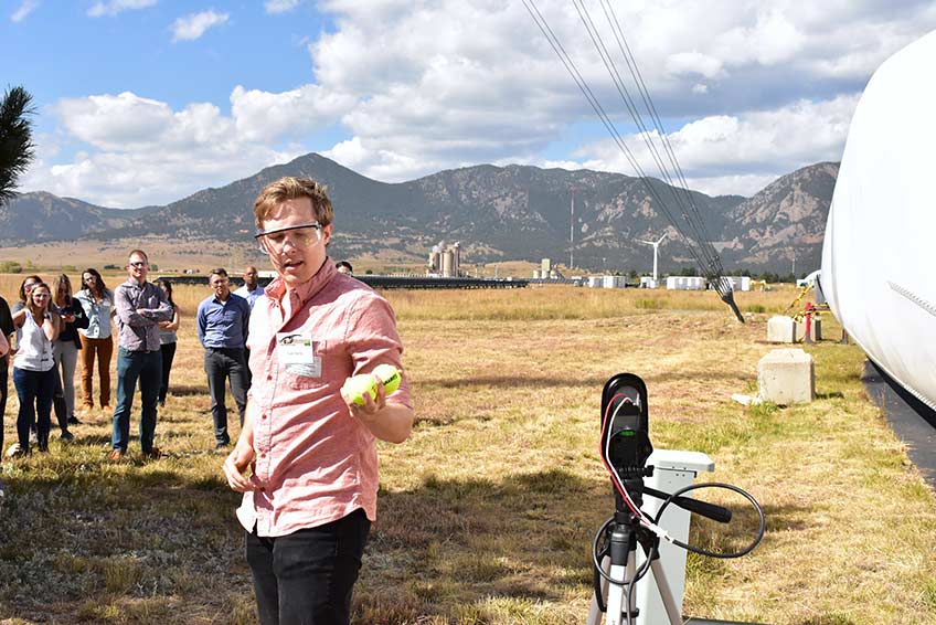 A man in a salmon-colored shirt holding two tennis balls demonstrates a wildlife impact detection technology to a group of onlookers.