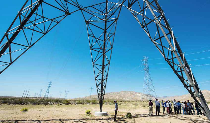 Group of people standing underneath electrical tower with one person in the middle looking straight up at it.