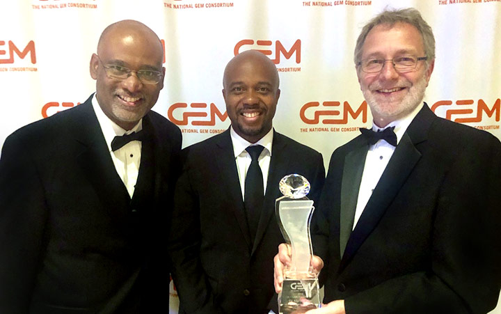 Photo of three men wearing tuxedos. One holds a glass trophy.