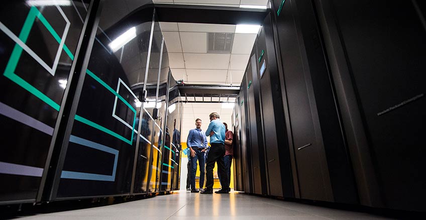 Three men stand between a row of computers in NREL’s High Performance Computer Data Center.