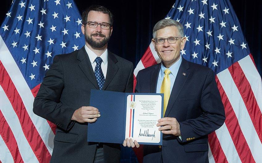 Two men shake hands in front of American flags.