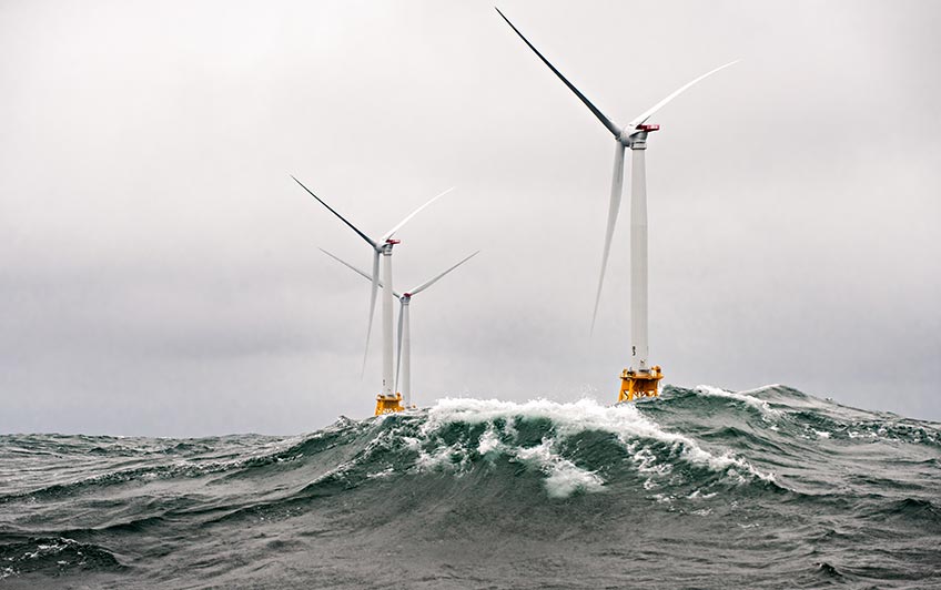 Beneath a stormy sky, three offshore wind turbines rise over high ocean waves.