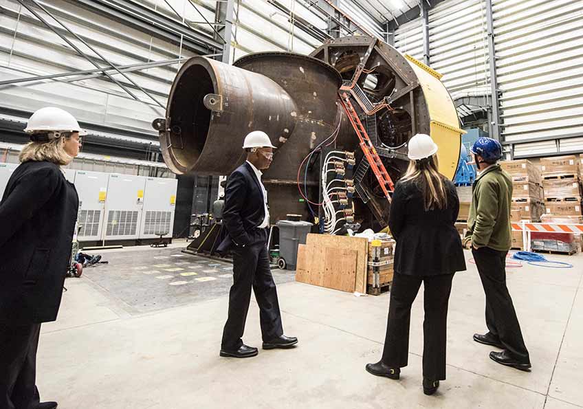 Four people in hard hats, inside a building, stand next to a large dynometer device. 
