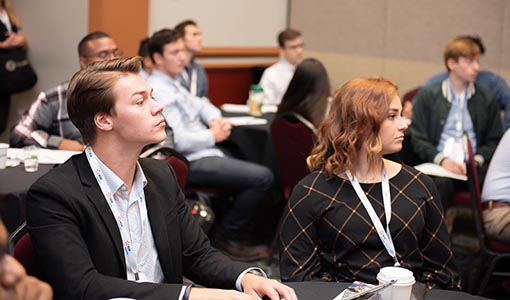 A young man and woman listen to a speaker.