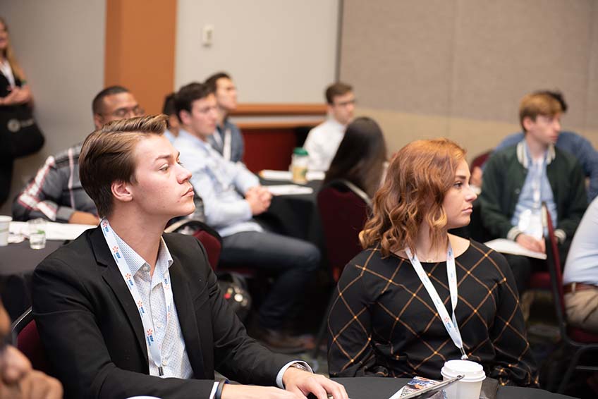 A young man and woman listen to a speaker.