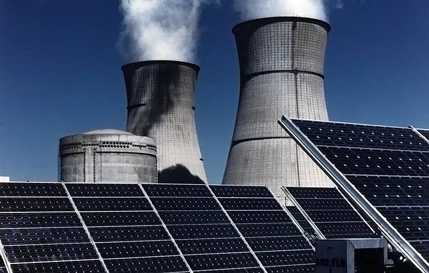 Photo of power plant cooling towers emitting water vapor with black solar panels in the foreground.