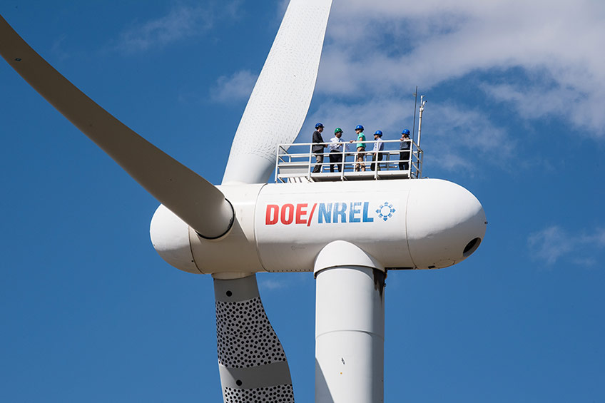 Photo of the top of a wind turbine, five people wearing hard hats are standing on top, blue skies with white clouds behind them. 