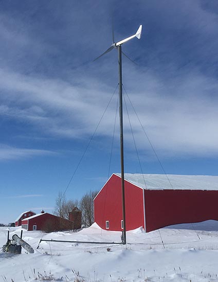Photo of a small wind turbine rising into the sky above a barn in a snowy field.