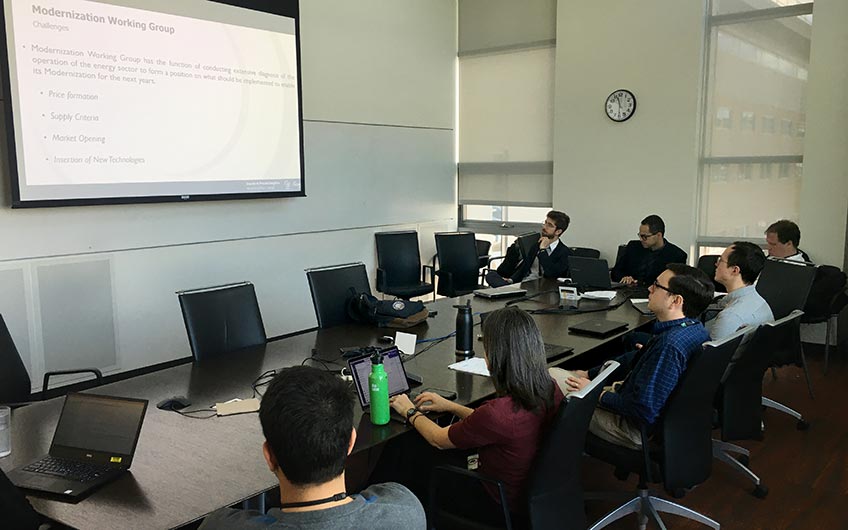 7 people sitting around a table looking at a large projection screen.