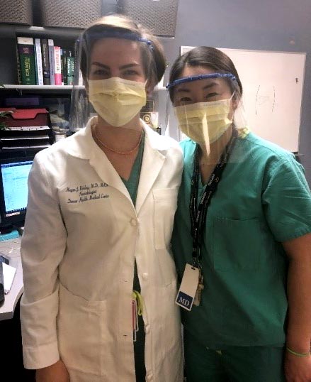 Two women pose for picture wearing medical uniforms and face shields.