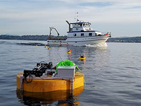Photo of a small wave energy converter floating in a body of water, buoys and a boat are in the background.