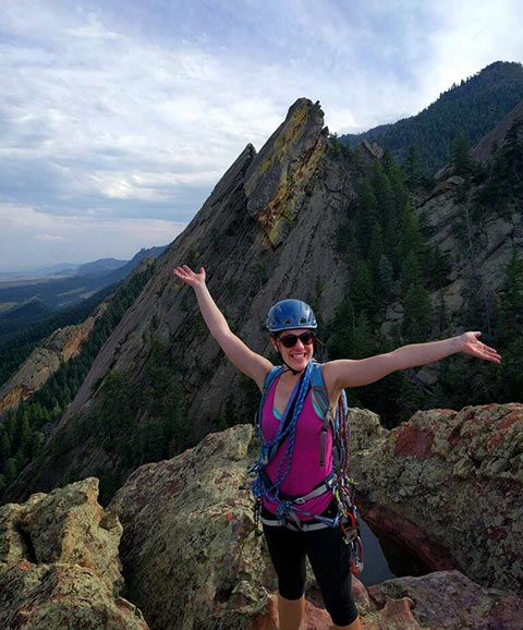 Photo of a woman rock climber at the top of a mountain with arms in the air after finishing a climb. Mountains in background.