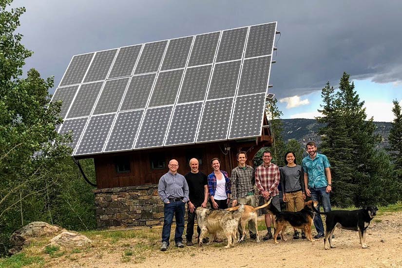 Photo of group of people with their dogs standing in front a cabin with big solar panels on the roof. Mountains in the background.