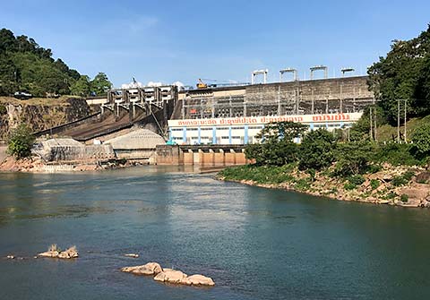 A hydropower dam in the Lao PDR with a blue sky above it and a body of water in the foreground.