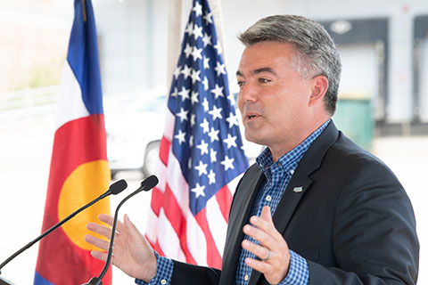 A man speaking at a podium with the U.S. flag and the Colorado flag in the background