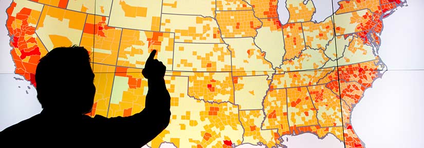 Photo of the back of an unidentified person pointing to a screen that shows a map of the United States. Different shades of orange on the map show varying levels of customer adoption of distributed energy resources. The coasts and cities have higher projected adoption.