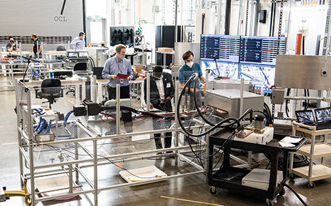 Three men stand next to large test benches with electric charging connectors plugged into inlets.