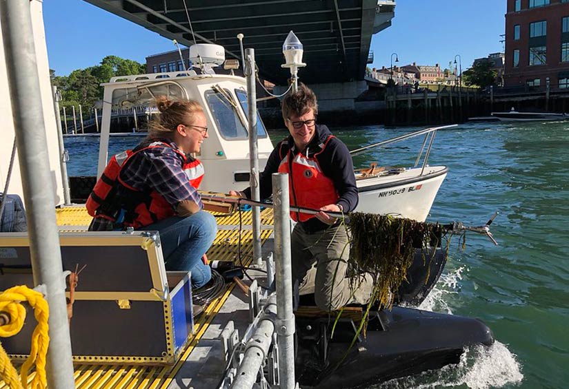 Photo of a woman and a man near a body of water, wearing lifejackets; a boat is behind them. The man is holding a long pole with seaweed on the end of it.