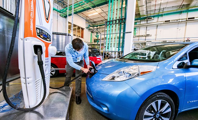 Image of an electric car inside a laboratory, charging station on the left-hand side of the photo, man is holding a charging line and placing it into a car.