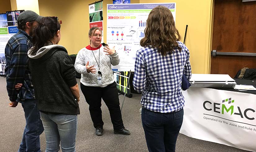 Photo of a group of three people standing and listening to a woman present in front of a poster. The CEMAC sign is in the foreground.