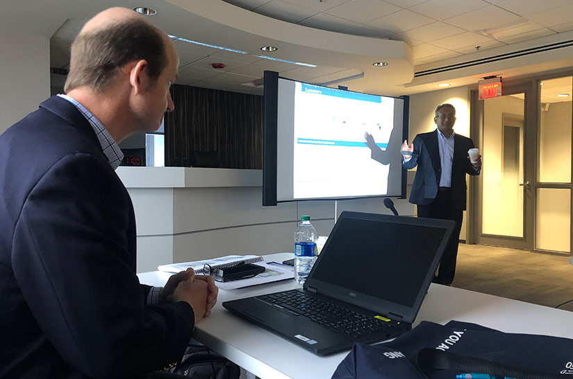 A man seated at a laptop watches a standing man present information on a projection screen