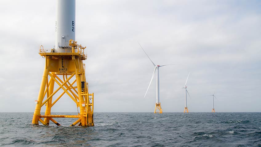 Photo of four wind turbines in the ocean against a gray sky.