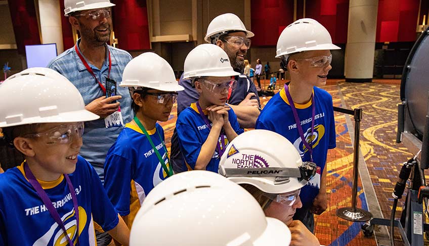 Photo of six children and two men wearing hard hats inside a banquet room, looking toward a piece of equipment on the right-hand side of the image. 