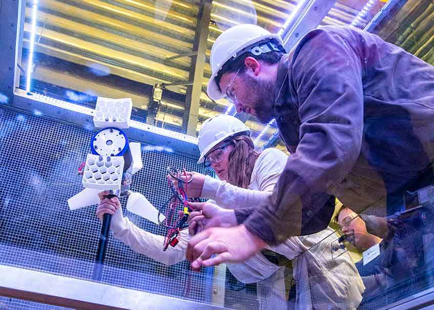 A young woman in a hard hat and her teacher work on a small wind turbine in a wind tunnel.