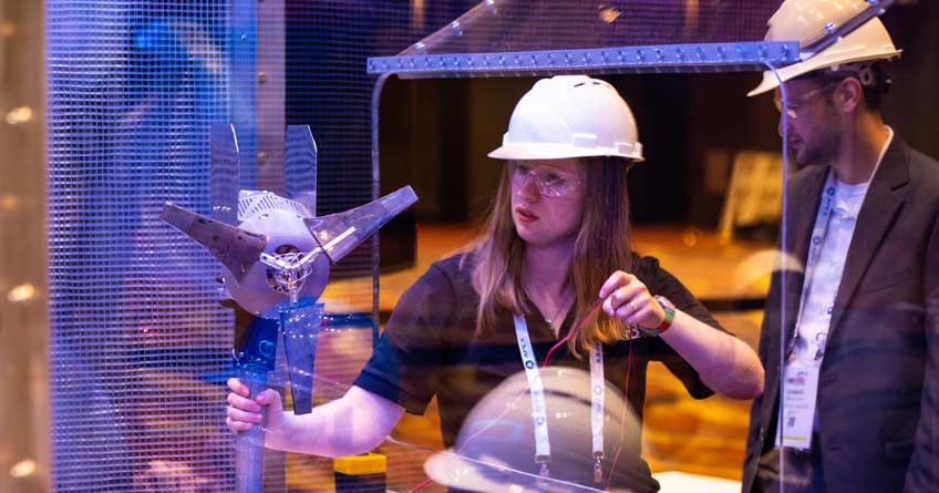 A young woman in a hard hat works on a small wind turbine in a wind tunnel.