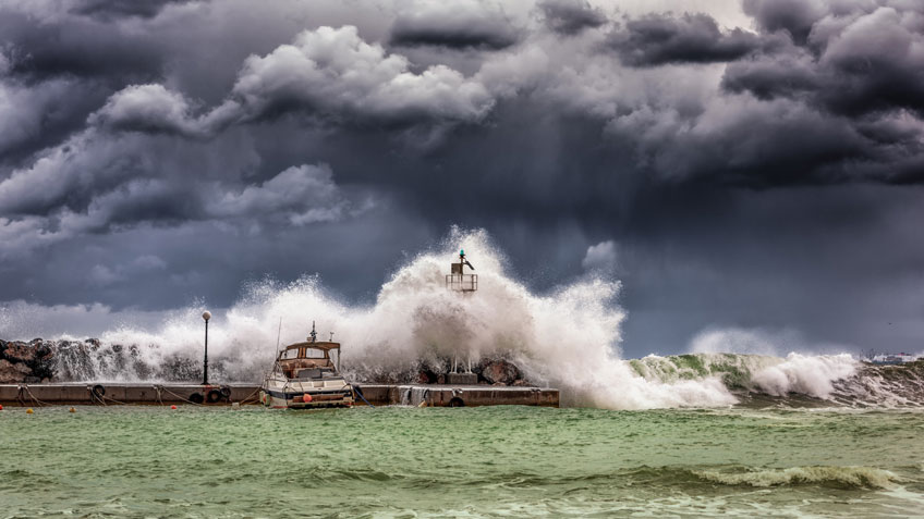 Photo of a boat a dock with stormy skies above and large waves crashing behind it.
