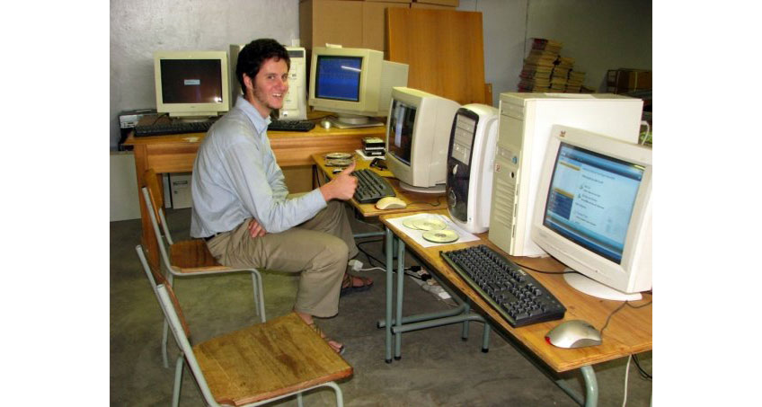 Photo of a young man sitting at a computer with a smile and giving a thumbs up. Several other computers surround him.