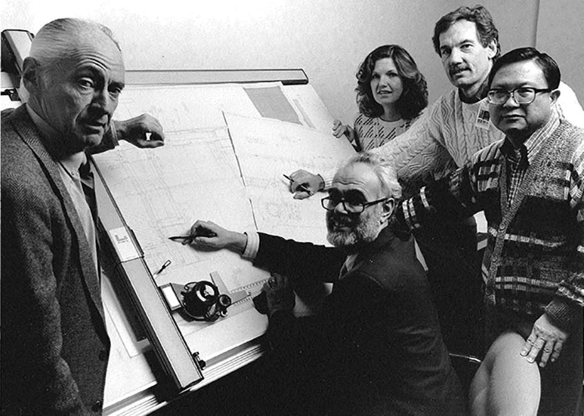 Black and white photograph of five people staged around a drawing board.
