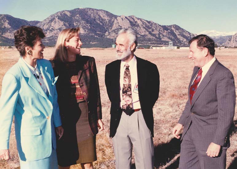 Two woman (left) and two men (right) stand in an open field with mountains in the background.