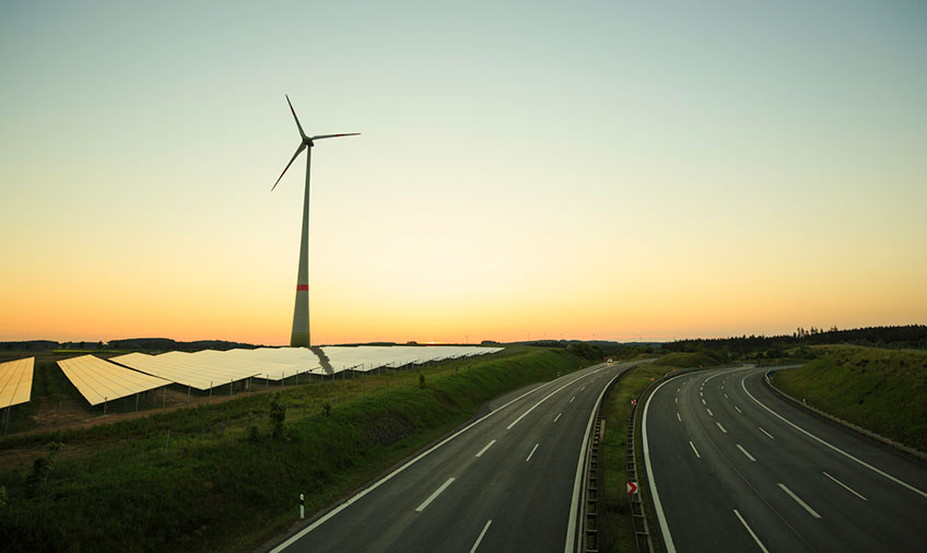 Photo at sunset of highway on the right and solar panels and a wind turbine on the left.