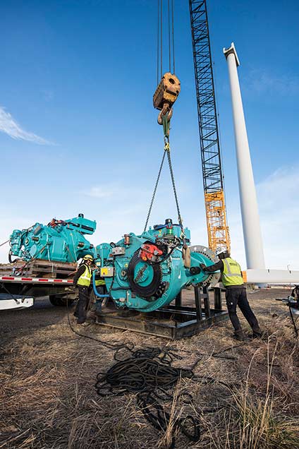 A crane lifts lowers a wind turbine gearbox onto a wooden pallet.