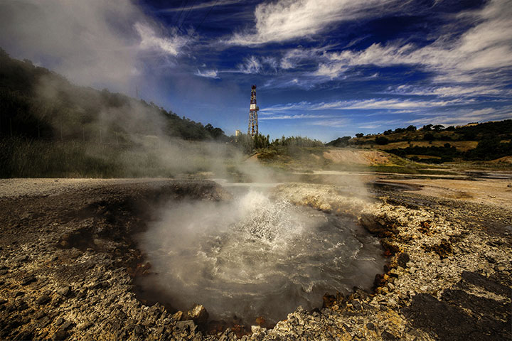 Photo of steamy, bubbling pool of water in outdoor setting with drilling rig in the background.