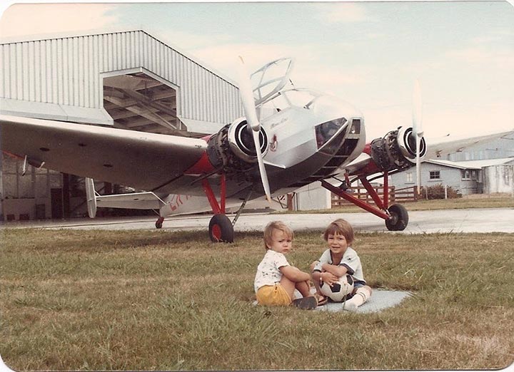 Photo of two young children sitting in grass in front of an airplane outside of a hangar.