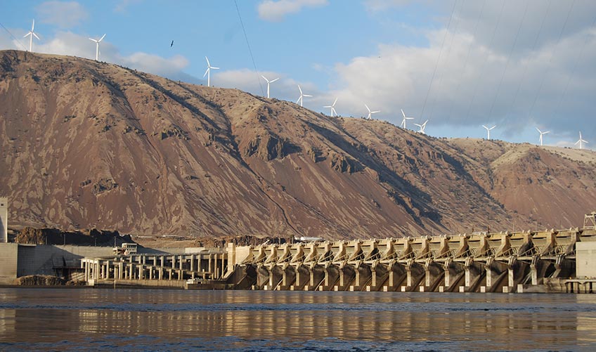 Photo of a hydropower plant with wind turbines situated atop mountains in the background.