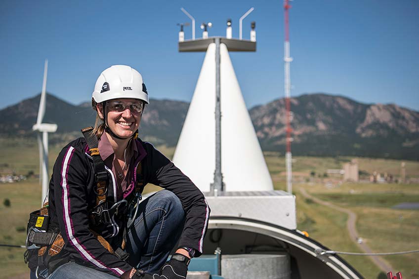 A female researcher with safety equipment crouches on top of a turbine nacelle with a mountain backdrop. 