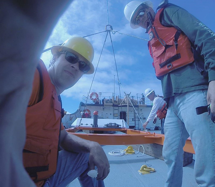 Photo of two people wearing hardhats, safety vests, and sunglasses on a boat. A third person is adjusting a cable in the background.