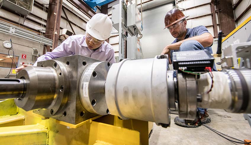 A photo of two men wearing hardhats crouching behind a metal testing device.