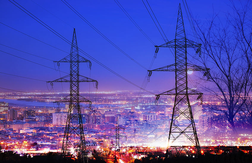 Photo of a lit-up cityscape at night with power transmission towers and power lines in the foreground.