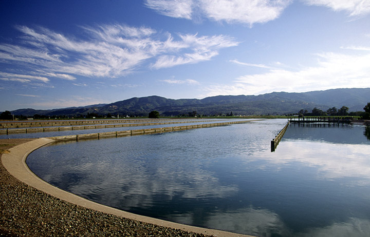 Photo of a large water resource recovery pond with clouds reflected in it, mountains in the background, and cloud-filled sky above.