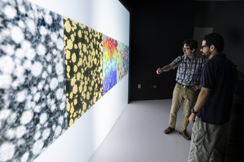 Two men stand in front of a giant screen with a black and yellow 3D rendering of a lithium-ion battery electrode. 