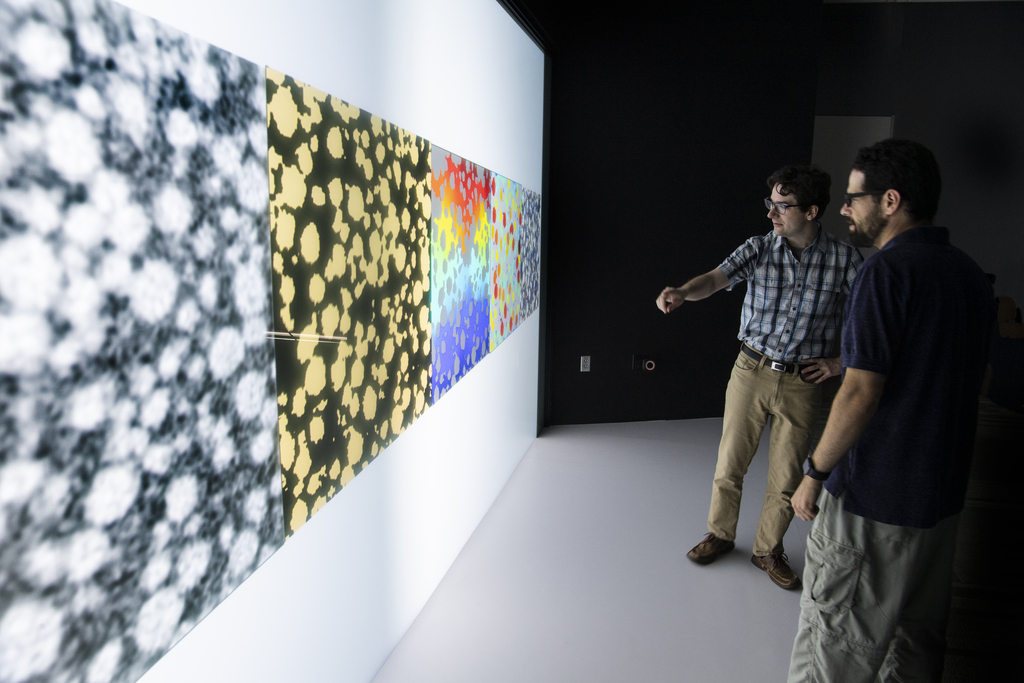 Two men stand in front of a giant screen with a black and yellow 3D rendering of a lithium-ion battery electrode.