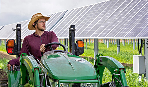 Photo of a man driving a tractor in a field next to solar panels