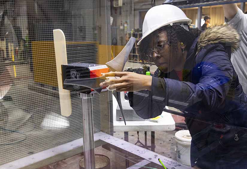 A young woman in a hard hat and safety glasses adjusts a small wind turbine.